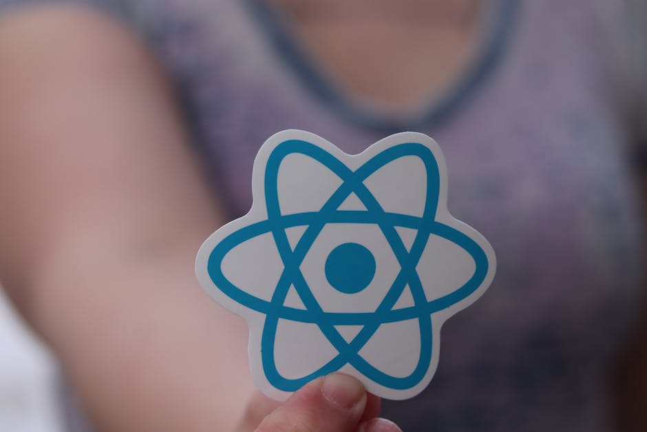 A person is holding a small, circular sticker with a blue atomic model symbol in the foreground, with a blurred background of a person wearing a gray shirt and purple top. The scene appears to be indo