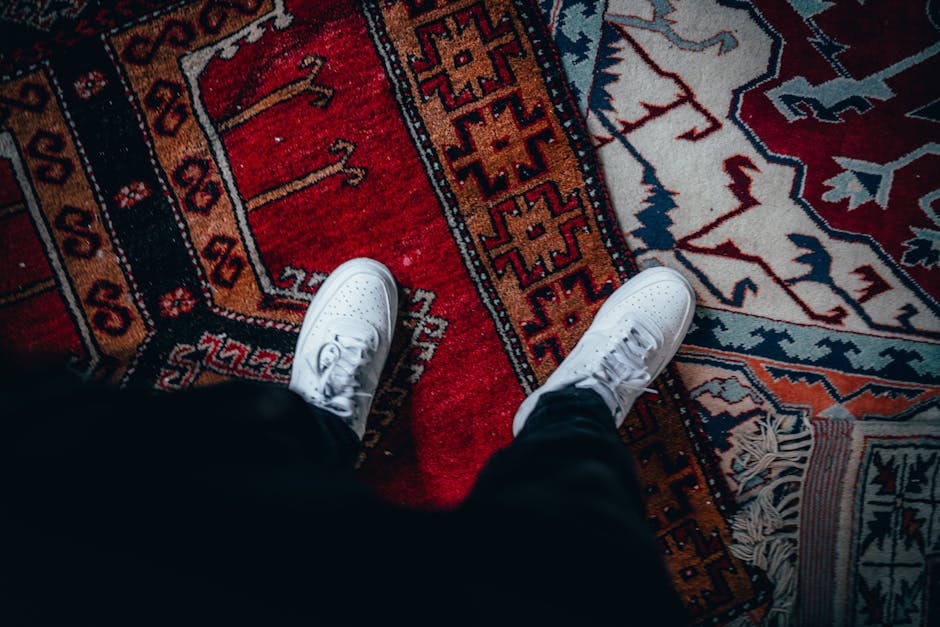 A top-down view of a person's white sneakers standing on a vibrant, patterned Oriental rug in a room with soft lighting. The rug features intricate geometric designs in red, blue, orange, and cream co